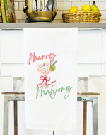 Load image into Gallery viewer, A white kitchen towel hanging on a metal rack in a bright kitchen, printed with the words 'Merry Mahjong' in red and green script around a small illustrated game board with red ribbon accents.
