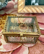 Load image into Gallery viewer, A small rectangular jewelry box with a clear glass top and ornate gold-toned metal sides, shown on a pink floral surface. A ring is placed inside the box.
